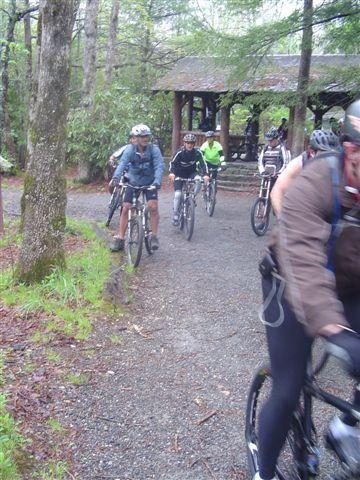 A group of mountain bikers riding along a winding gravel path in a wooded area. The scene is lush with greenery and includes a rustic shelter in the background. Some riders are wearing helmets and biking gear while others are dressed in casual attire, indicating a mix of skill levels. The atmosphere appears adventurous and active. Black Mountain mountain bike trail.