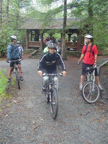 A group of four mountain bikers is gathered on a gravel path in a wooded area. One cyclist is in motion on a bike, while the others are either standing or preparing to ride. A wooden shelter is visible in the background, surrounded by trees. The atmosphere suggests an outdoor adventure or biking excursion. Black Mountain mountain bike trail.