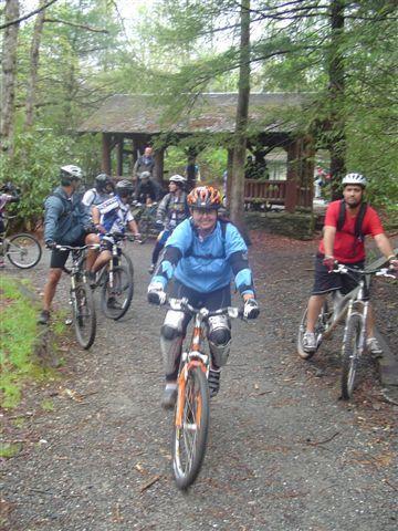 A group of mountain bikers riding along a gravel path in a forested area, with a covered shelter visible in the background. The cyclists are dressed in various biking gear, including helmets and protective clothing, as they navigate through the lush greenery. Black Mountain mountain bike trail.