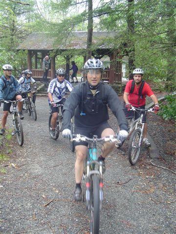 A group of mountain bikers riding on a gravel path through a wooded area. They are wearing helmets and biking gear, with some riders approaching a gazebo in the background. Black Mountain mountain bike trail.