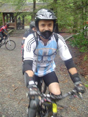 A person in a cycling jersey featuring the colors of Argentina rides a mountain bike on a gravel path surrounded by trees. They are wearing a helmet and protective gear, with a mask covering their mouth and nose. In the background, another cyclist can be seen, and a wooden structure is visible among the greenery. Black Mountain mountain bike trail.