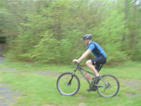 A person riding a mountain bike on a grassy path surrounded by greenery, with a blurred motion effect suggesting speed. The cyclist is wearing a blue and black jersey and a helmet. Black Mountain mountain bike trail.
