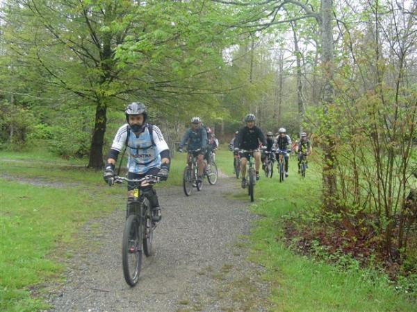 A group of mountain bikers riding on a gravel path through a green, wooded area. The scene is slightly rainy, with trees in the background and lush foliage on either side of the trail. The riders are wearing helmets and cycling gear, and some are positioned in a line, showcasing a variety of biking styles. Black Mountain mountain bike trail.