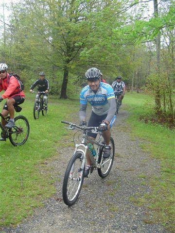 A group of four mountain bikers riding on a gravel path through a wooded area. In the foreground, a cyclist in a blue and white jersey is actively biking towards the camera, while three other cyclists are seen in the background, each wearing different colored jerseys and helmets. The scene is set in a lush, green environment, indicating an outdoor activity in nature. Black Mountain mountain bike trail.