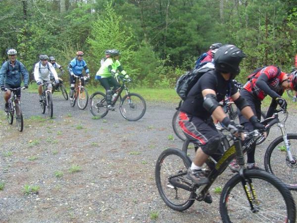 A group of mountain bikers wearing helmets and protective gear, riding on a gravel path surrounded by greenery. The cyclists are in various stages of riding, with some focusing on the trail ahead while others adjust their gear. The scene captures the energy and camaraderie of an outdoor biking event. Black Mountain mountain bike trail.