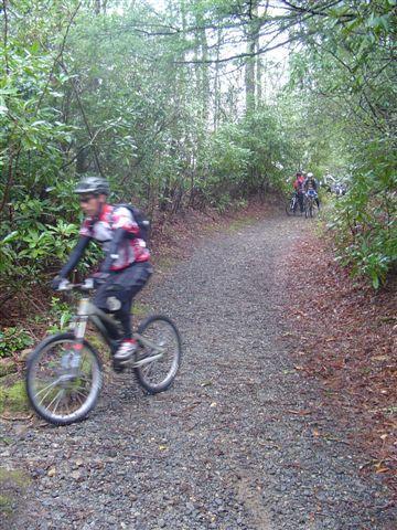 A person riding a mountain bike on a gravel trail surrounded by greenery. In the background, other cyclists can be seen on the same path. The environment appears lush and forested, suggesting an outdoor recreational area. Black Mountain mountain bike trail.