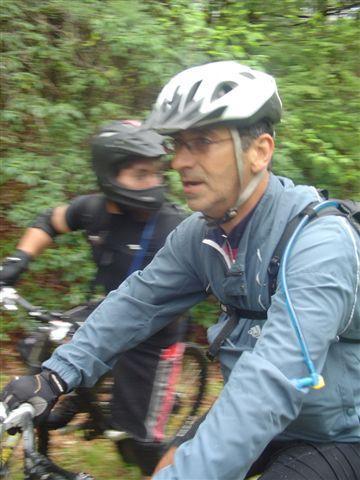 Two bicyclists riding on a wooded trail. One cyclist, wearing a helmet and a blue jacket, is in focus, while the second cyclist, dressed in black with a helmet, appears slightly blurred in the background, indicating motion. The surrounding environment is lush with greenery. Black Mountain mountain bike trail.