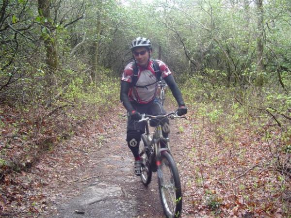 A mountain biker riding on a narrow, leaf-covered trail through a lush, green forest. The cyclist is wearing a black helmet, a red and white cycling jersey, and padded shorts. Surrounding vegetation includes trees and shrubs, creating a natural, outdoor setting. Black Mountain mountain bike trail.