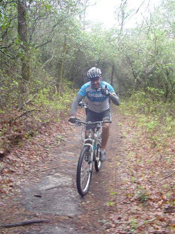 A mountain biker riding along a narrow trail surrounded by dense greenery and trees, wearing a helmet and cycling gear, and gesturing with one hand. Black Mountain mountain bike trail.