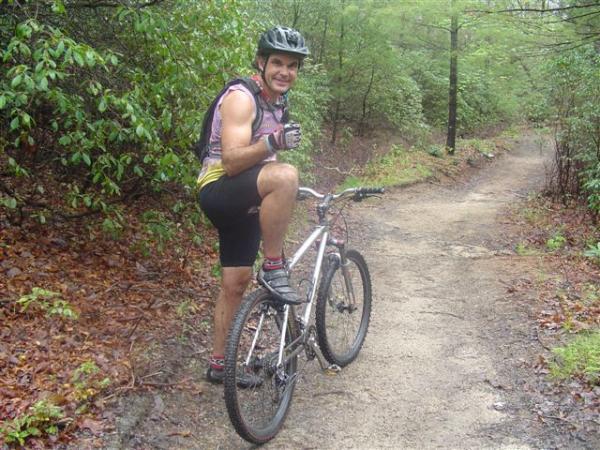 A male mountain biker gives a thumbs-up while sitting on his bike on a dirt trail surrounded by greenery. He is wearing a helmet and sporty biking attire, showcasing a confident and enthusiastic expression. The scene captures a moment of outdoor adventure in a wooded area, with a path winding through the foliage. Black Mountain mountain bike trail.