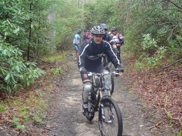 A group of mountain bikers riding along a narrow trail in a wooded area, surrounded by greenery. The leading rider is wearing a helmet, gloves, and protective gear, and is focused on the path ahead. Other cyclists can be seen in the background, also navigating the trail. The terrain appears slightly muddy, indicating recent rain. Black Mountain mountain bike trail.