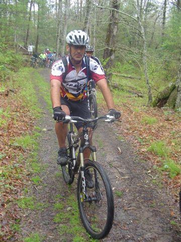 A man wearing a helmet and cycling gear rides a mountain bike along a muddy trail in a forested area, surrounded by trees and greenery. Black Mountain mountain bike trail.