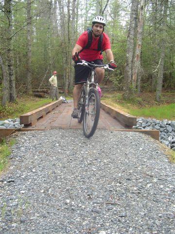 A mountain biker wearing a helmet and a red shirt rides over a wooden bridge on a gravel path surrounded by trees. In the background, a person is visible walking on the trail. Black Mountain mountain bike trail.