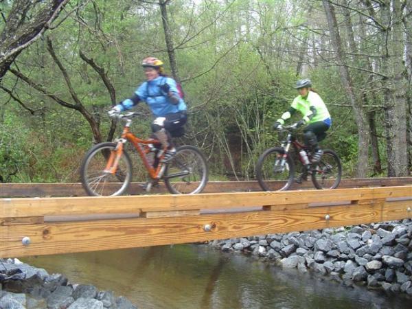 Two mountain bikers ride across a wooden bridge over a stream in a lush, green forest. One cyclist wears a blue jacket and helmet, while the other is dressed in a bright green jacket. The scene captures the hikers' movement as they navigate the bridge, surrounded by trees and rocky terrain. Black Mountain mountain bike trail.
