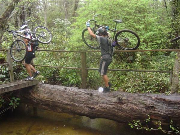 Two cyclists are crossing a log bridge in a wooded area while carrying their bicycles. The scene is lush with greenery and trees in the background, and the ground below shows a small stream. One cyclist is in a colorful jersey and helmet, while the other is wearing a more muted outfit. Both are focused on navigating the challenging terrain. Black Mountain mountain bike trail.