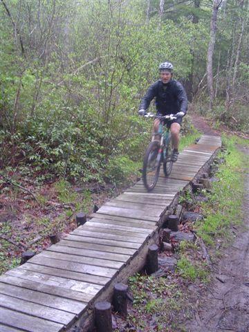 A cyclist riding a mountain bike along a narrow wooden boardwalk trail surrounded by lush greenery in a forested area. The weather appears overcast, adding a misty atmosphere to the scene. Black Mountain mountain bike trail.