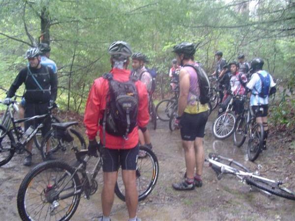 A group of mountain bikers gathered on a muddy trail during a rainy day, wearing helmets and cycling gear. Some riders are on their bikes while others are engaged in conversation, surrounded by dense greenery in a forested area. Black Mountain mountain bike trail.