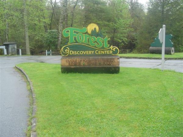Sign for the Forest Discovery Center, featuring vibrant green lettering on a wooden background, surrounded by lush grass and trees. The sign includes the phrase "Cradle of Forestry in America." The area appears quiet and serene, with a pathway leading to the center. Black Mountain mountain bike trail.