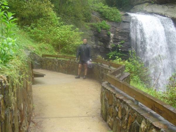 A person standing on a paved path near a waterfall, surrounded by lush greenery. The pathway features a stone railing, and the individual is dressed in outdoor attire. Water cascades over rocky ledges in the background. Black Mountain mountain bike trail.