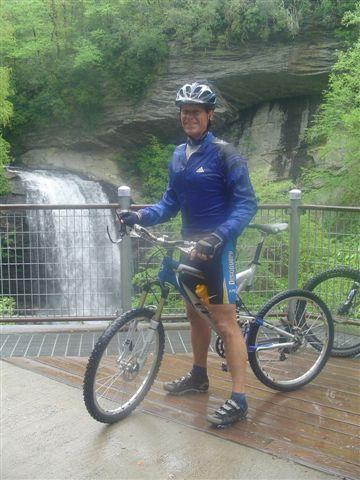 A person wearing a blue cycling jersey and helmet stands next to a mountain bike on a wooden platform overlooking a waterfall surrounded by lush greenery. Black Mountain mountain bike trail.