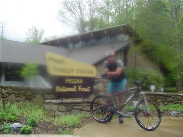 A cyclist in casual biking attire stands next to a sign for the Pisgah Ranger Station, located at Pisgah National Forest. The background features the ranger station building, surrounded by greenery. The image appears slightly blurred due to fog or mist. Black Mountain mountain bike trail.