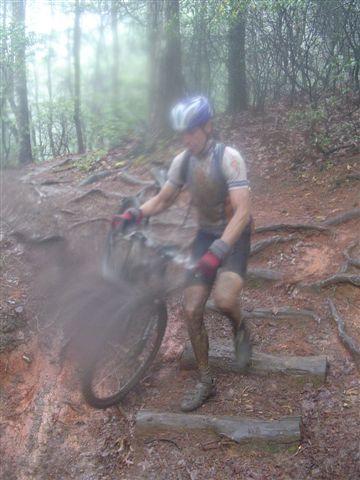 A mountain biker navigating a muddy trail surrounded by trees, appearing partially obscured by mist. The rider is wearing a helmet and gloves, and is attempting to lift the bike over wooden steps that are integrated into the rugged terrain. The scene conveys a challenging outdoor environment. Black Mountain mountain bike trail.