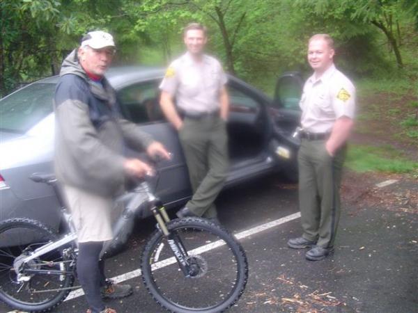 A blurry image of a man in cycling attire standing next to a mountain bike, conversing with two park rangers in uniforms near a parked car in a wooded area. The scene is set in a rainy environment with lush greenery in the background. Black Mountain mountain bike trail.