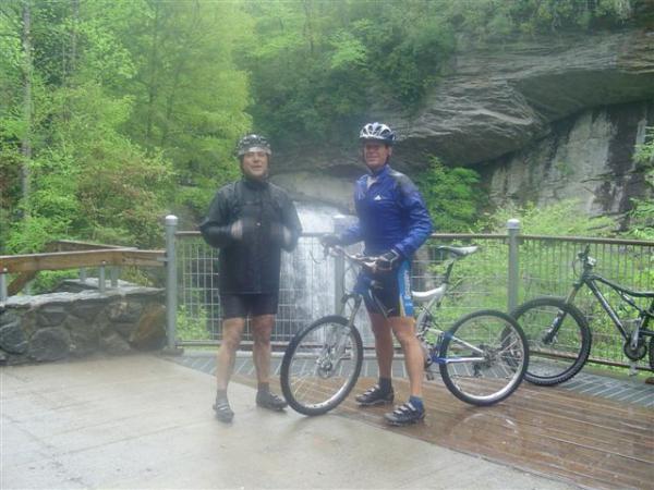 Two cyclists stand near a waterfall, surrounded by lush greenery. One cyclist wears a black jacket and helmet, while the other is dressed in a blue cycling outfit. Both bikes are parked beside them on a wooden platform. The scene captures a rainy day in a scenic outdoor setting. Black Mountain mountain bike trail.