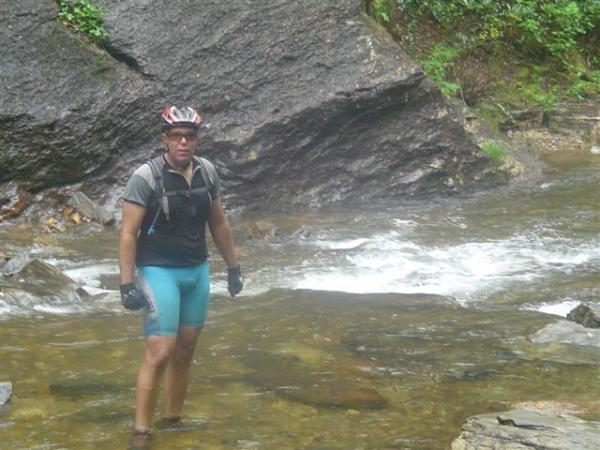 A person wearing cycling gear and a helmet is wading through a shallow stream surrounded by rocks and greenery. The water is flowing gently around them, and they appear focused on navigating the terrain. Black Mountain mountain bike trail.
