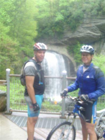 Two cyclists stand in front of a waterfall, wearing helmets and cycling gear. They are posed near their mountain bikes, with lush greenery surrounding them and the cascading water visible in the background. The setting appears to be a scenic outdoor location, showcasing nature. Black Mountain mountain bike trail.