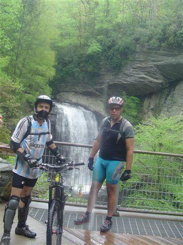 Two cyclists stand next to a mountain bike at a scenic waterfall in a lush, green forest. One cyclist wears a helmet, knee and elbow pads, and a jersey featuring the word "Argentina," while the other sports a helmet and cycling shorts. The waterfall cascades down rocky cliffs in the background, surrounded by vibrant foliage. Black Mountain mountain bike trail.