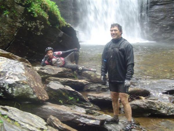 Two individuals stand amidst rocky terrain near a waterfall. One man is smiling and wearing a black cycling outfit, while another is reclining on the rocks, partially obscured, wearing a helmet and casual clothing. They appear to be enjoying the natural surroundings, with water flowing in the background and greenery framing the scene. Black Mountain mountain bike trail.