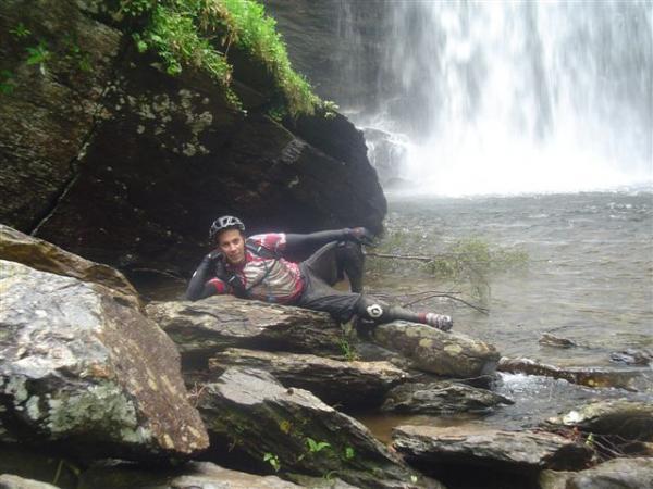 A person in biking gear relaxes on a rock near a waterfall, surrounded by lush greenery and water cascading in the background. Black Mountain mountain bike trail.
