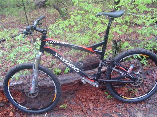 A black and orange specialized mountain bike is parked on a forest floor surrounded by greenery. The bike features thick tires, a sturdy frame, and a shock absorber on the front fork, suggesting it's designed for rugged terrain. Black Mountain mountain bike trail.