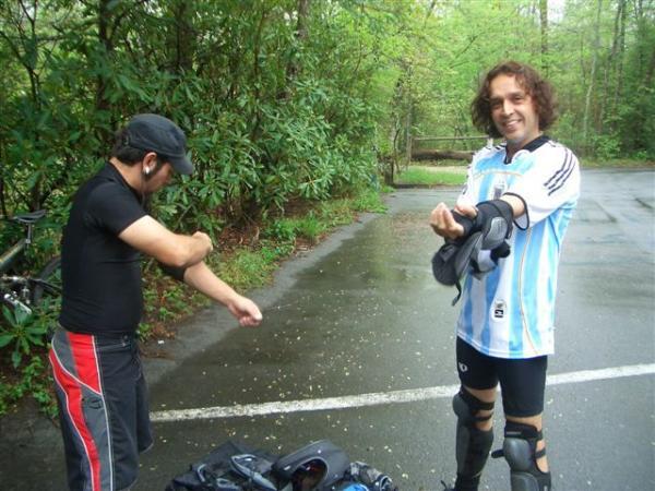 Two individuals preparing for an outdoor activity on a rainy day. One person, wearing a black shirt and cap, is putting on protective gear. The other, dressed in a blue and white jersey, is smiling while adjusting his wrist guard. They are surrounded by lush greenery along a paved road. Black Mountain mountain bike trail.