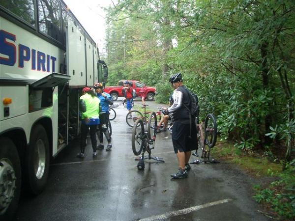 A group of cyclists in rain gear prepare their bicycles near a large bus on a wet, wooded road. Some bikes are positioned upside down for adjustments, while others are being handled by riders. A red truck is visible in the background amidst greenery. Black Mountain mountain bike trail.