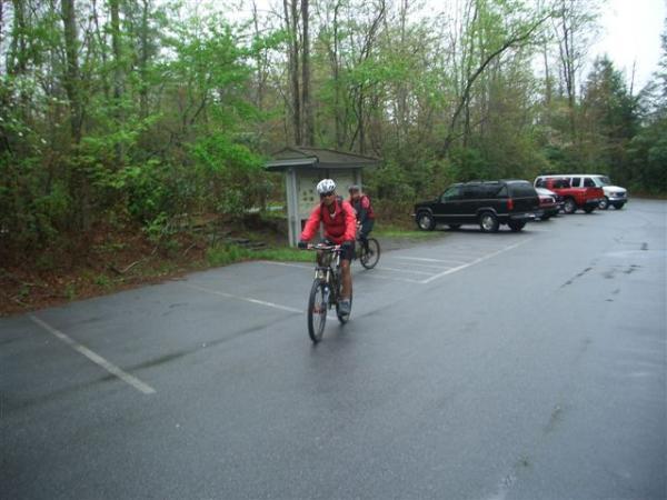 Two cyclists in bright red jackets ride through a wet parking lot surrounded by trees. The scene captures the cyclists approaching a small shelter, with parked vehicles visible in the background. The atmosphere suggests a rainy day in a natural setting. Black Mountain mountain bike trail.