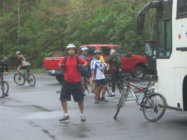A group of cyclists preparing for a ride in a parking area surrounded by trees. One man in a red t-shirt and helmet stands in the foreground, while others are seen near bicycles and a bus. Various bicycles are parked, and a red pickup truck is visible in the background. The atmosphere suggests an outdoor biking adventure. Black Mountain mountain bike trail.