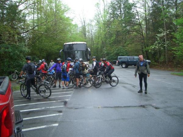 A group of mountain bikers gathers around their bicycles in a parking lot, with a large bus parked nearby. Trees and greenery can be seen in the background, indicating a natural setting. Some cyclists are wearing helmets and biking gear, while one individual stands off to the side, dressed differently. The ground appears wet, suggesting recent rain. Black Mountain mountain bike trail.