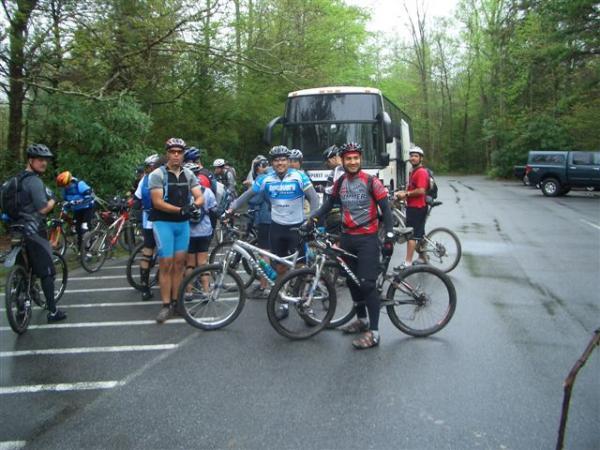 A group of cyclists wearing helmets and cycling gear are gathered in a parking lot surrounded by trees. They are standing next to their mountain bikes, with a large bus parked in the background. The scene appears to be set on a cloudy day, suggesting they are preparing for an outdoor biking adventure. Black Mountain mountain bike trail.