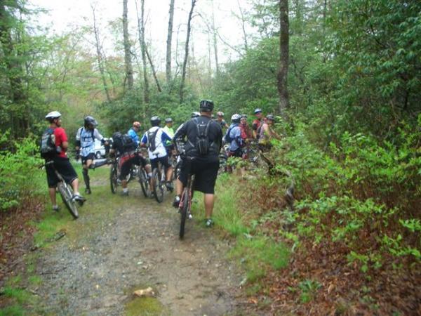 A group of mountain bikers gathered on a dirt path in a wooded area, surrounded by trees and greenery. Some riders are adjusting their bikes while others stand in conversation, all wearing helmets and cycling gear. The setting appears slightly damp, suggesting recent rain. Black Mountain mountain bike trail.