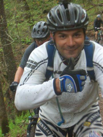 A group of mountain bikers in a wooded area. One rider in the foreground is smiling and giving a thumbs up, wearing a helmet and protective gear, while another cyclist is visible behind him. The trail is surrounded by greenery, indicating an outdoor adventure. Black Mountain mountain bike trail.