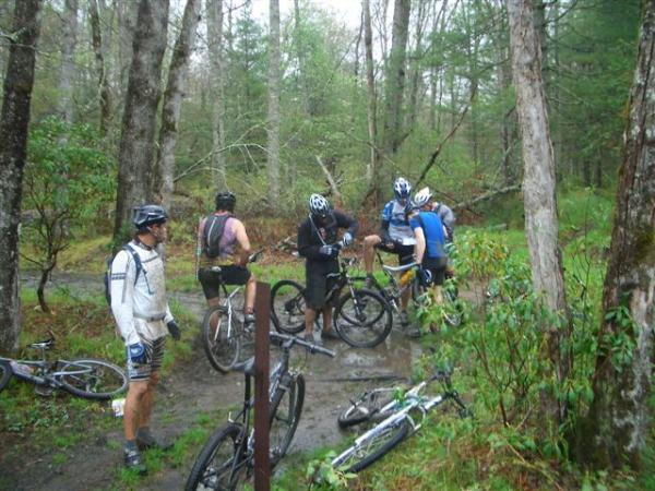 A group of five mountain bikers is gathered in a muddy area surrounded by trees in a forest. Some of the bikers are checking their bikes, while others appear to be resting. Several bikes are lying on the ground. The setting is rainy and lush with greenery, indicating an outdoor adventure. Black Mountain mountain bike trail.