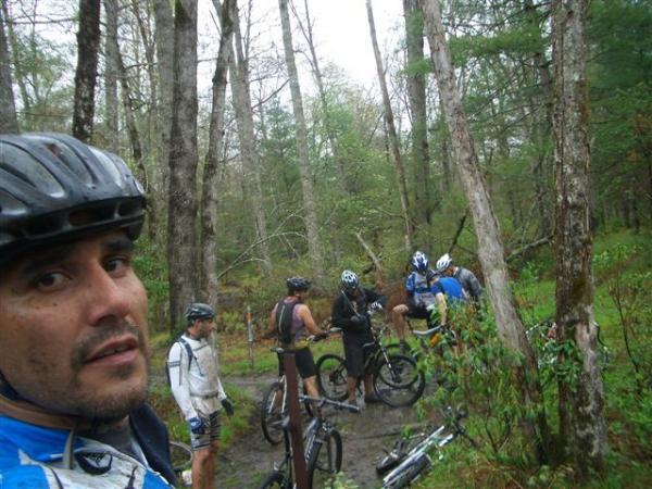 A group of mountain bikers pauses in a lush, wooded area during an outdoor ride. One cyclist in the foreground, wearing a helmet and a blue jersey, looks toward the camera, while several others are seen in the background, chatting and resting beside their bicycles. The scene is set in a rainy environment, with trees and greenery surrounding the group. Black Mountain mountain bike trail.
