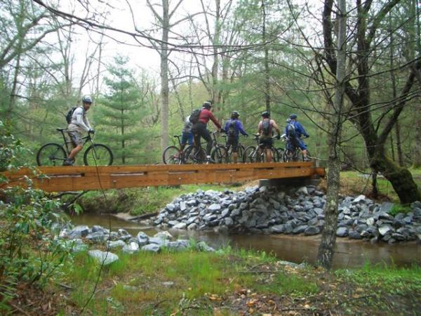 A group of six cyclists riding their mountain bikes across a wooden bridge over a small creek, surrounded by a lush green forest. The scene is set on a rainy day, with trees in the background and rocky terrain near the water's edge. Black Mountain mountain bike trail.