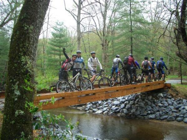 A group of mountain bikers poses on a wooden bridge over a stream in a forested setting. The riders are wearing helmets and biking gear, smiling and showing enthusiasm, with some raising their arms in excitement. Surrounding them are trees and greenery, indicating a natural outdoor environment. Black Mountain mountain bike trail.
