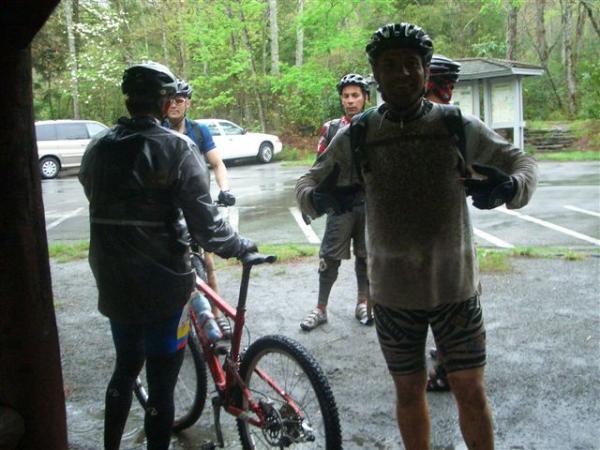 A group of four mountain bikers stands near a bike in the rain, with trees in the background. One rider is displaying a playful pose, showcasing their muddy attire. The others are dressed in cycling gear and helmets, ready to ride. The scene captures a rainy day adventure in a natural setting. Black Mountain mountain bike trail.