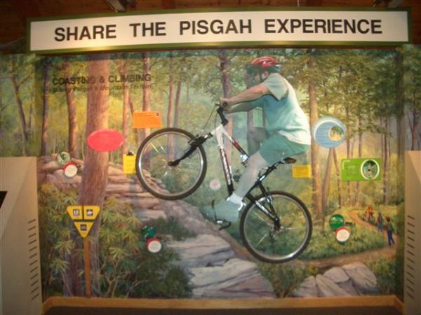 A man riding a mountain bike in mid-air, depicted on a mural in a forest setting, surrounded by informational signs about biking and climbing in Pisgah National Forest. The mural features vibrant greenery and a sense of adventure, inviting viewers to explore outdoor activities. The title "SHARE THE PISGAH EXPERIENCE" is prominently displayed above the mural. Black Mountain mountain bike trail.