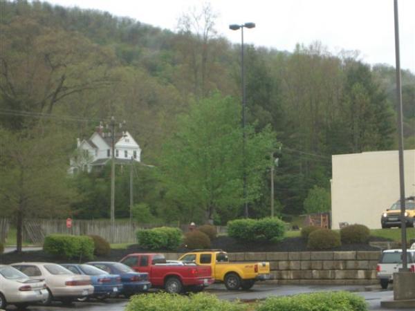 A view of a parking lot with several vehicles, including a red truck, a blue car, and a yellow truck. In the background, there is a lush green tree and hills covered with trees. A white house is partially visible on the left side of the image, and a beige building is seen on the right. The sky appears overcast, suggesting possibly rainy weather. Black Mountain mountain bike trail.