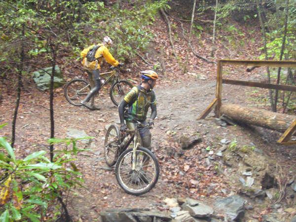 Two mountain bikers navigating a muddy trail in a wooded area. One rider is in a yellow jacket and helmet, while the other is wearing a green and black outfit. They are both pushing their bikes over rough terrain, with fallen leaves and rocks around them. A wooden barrier is visible on the path ahead. Black Mountain mountain bike trail.